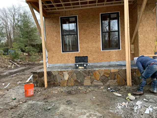Construction site with a worker placing stone veneer on the base of a partially finished house.