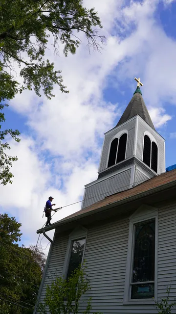 A harnessed roofer working on the roof of a white church below its steeple