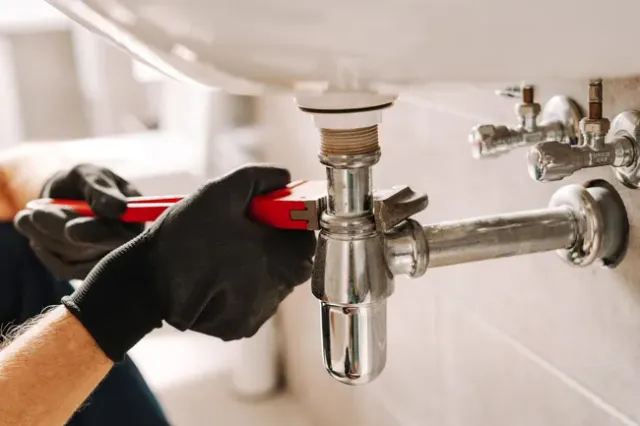 Plumber using a red pipe wrench to tighten a metal drain pipe under a white bathroom sink.