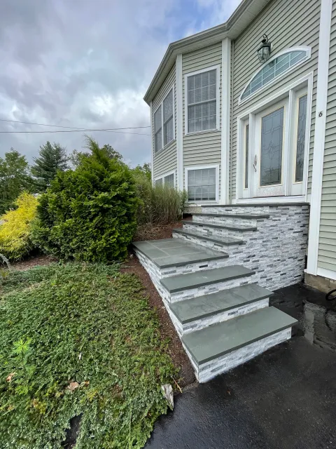 Exterior view of a house showing a front porch with grey and white stone stairs, green siding, and a white front door with stained glass, surrounded by landscaping and a wet black asphalt driveway.