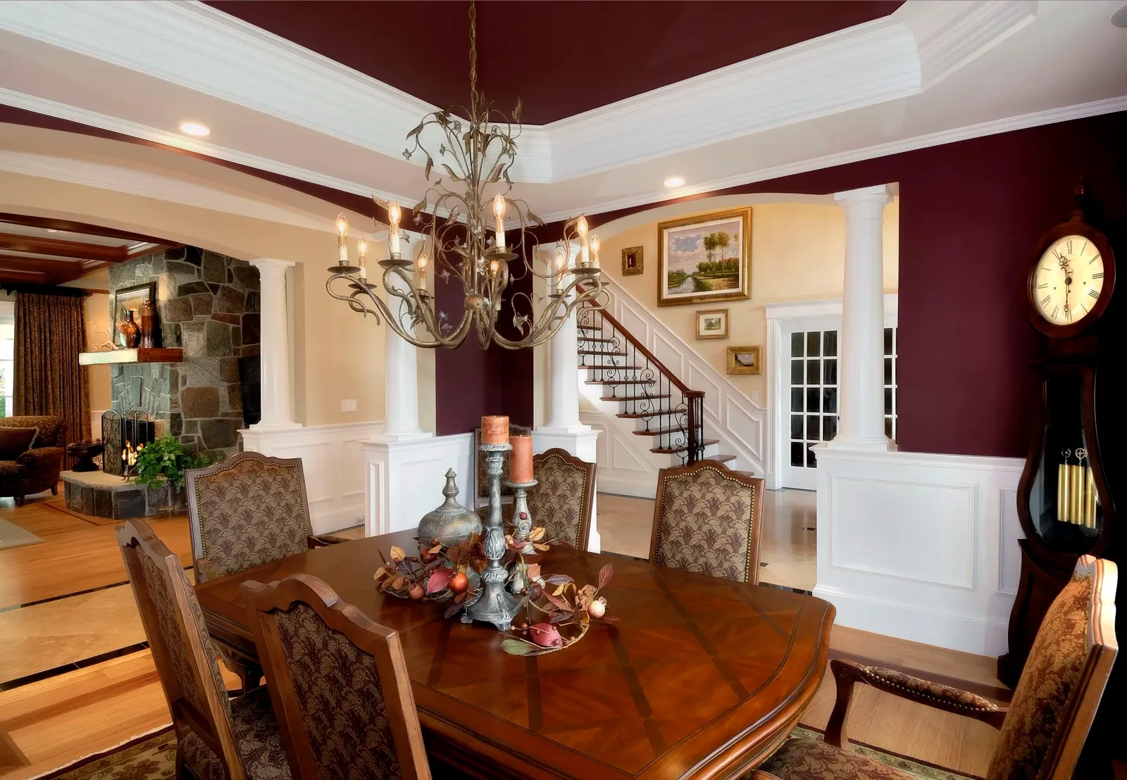 Interior of a dining room with a wooden table and chairs, a chandelier, and decorative items. The space features white wainscoting, a staircase with a dark wood handrail, and framed paintings on the wall. A grandfather clock is visible on the right s