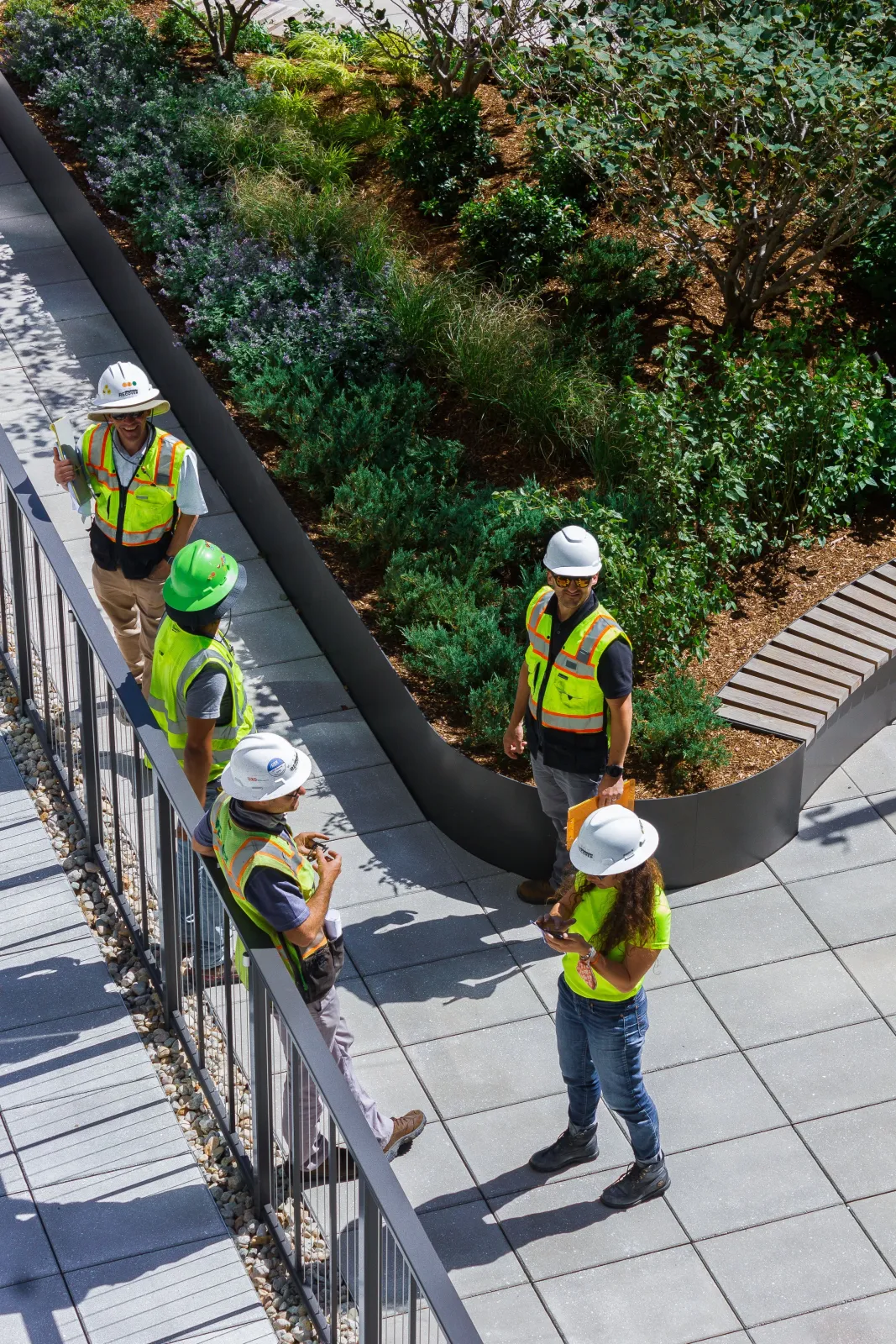 A group of workers and engineers wearing safety vests and hard hats walking and discussing on a rooftop garden with greenery and trees, some using their phones.