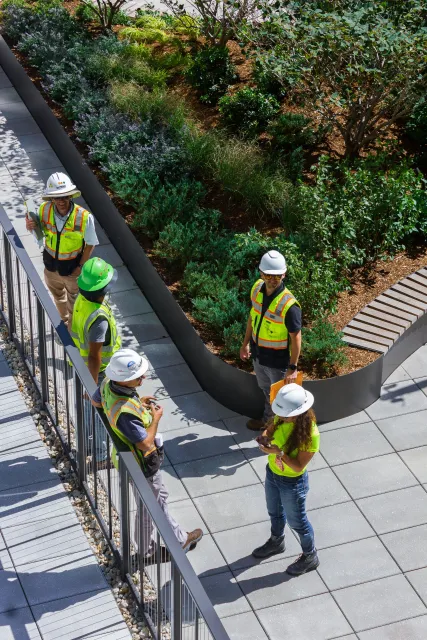 A group of workers and engineers wearing safety vests and hard hats walking and discussing on a rooftop garden with greenery and trees, some using their phones.
