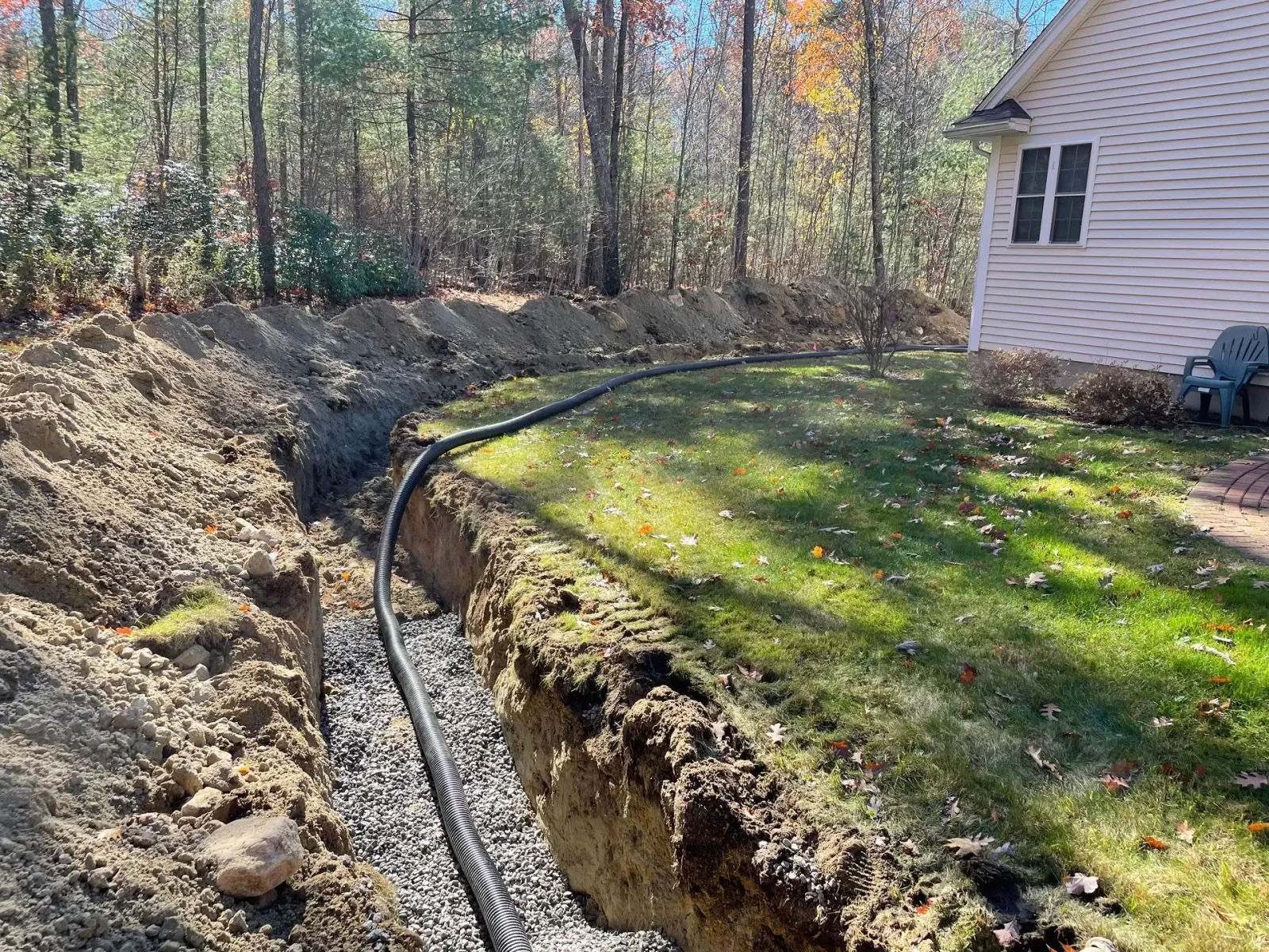 A black pipe in a trench with gravel, next to grass and a house, outdoors in daylight.