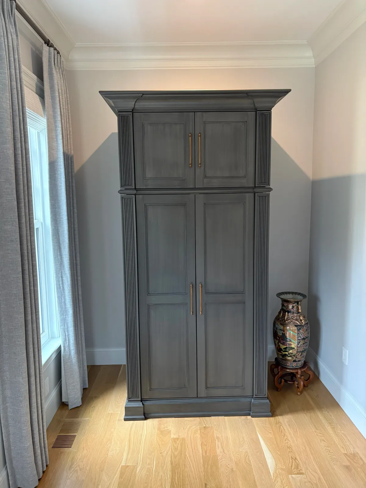 A tall, dark gray wooden cabinet with decorative molding and gold handles, positioned in a room with white walls, crown molding, and hardwood floors. To the right of the cabinet is a decorative Chinese vase on a carved wooden stand, and to the left a