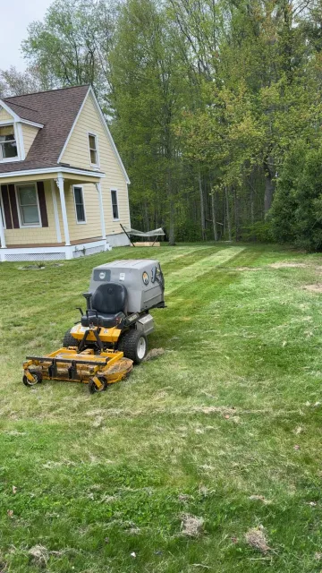 A riding lawn mower on a grassy yard beside a yellow house and trees.