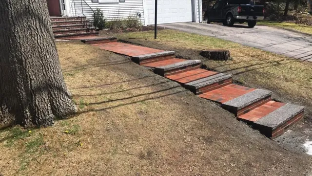 A set of brick stairs with concrete edges leading from a sidewalk to a house porch, with a large tree on the left and a driveway on the right.