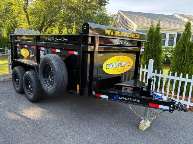 Black utility trailer with spare tires and yellow logos parked beside a white picket fence