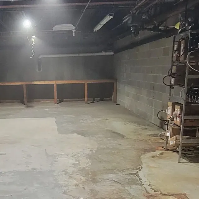 Empty basement or garage with concrete floor, cinder block wall, and metal shelving with boxed items on the right side. Overhead industrial lighting and exposed ceiling pipes.
