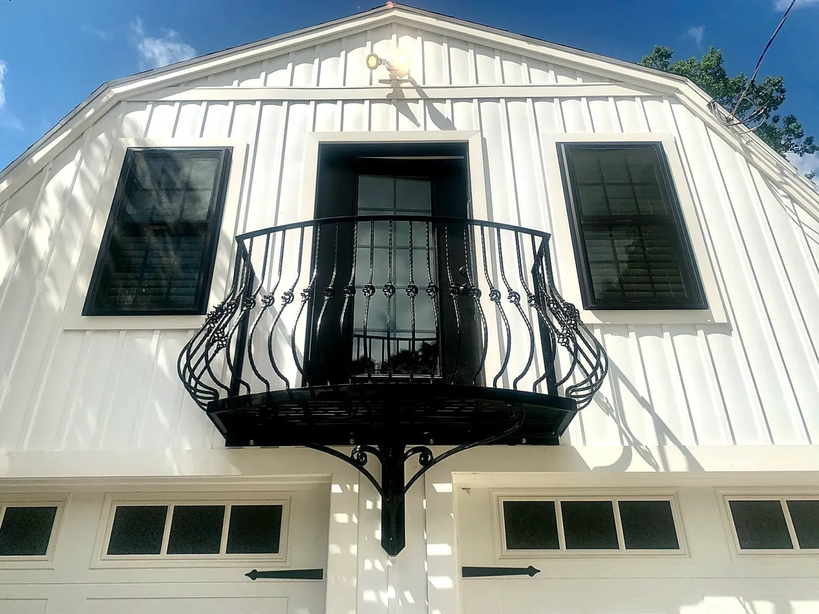 A white house with a black wrought iron balcony on the second floor, set against a blue sky with some clouds. The house has large black framed windows and a gabled roof. There is a black outdoor light fixture above the balcony.