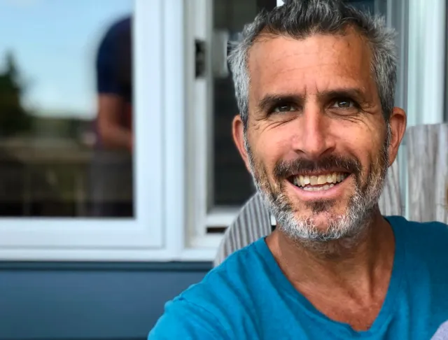 A smiling middle-aged man with gray hair and a beard, wearing a blue shirt, sitting outdoors near a window with a reflection of a person in the glass.