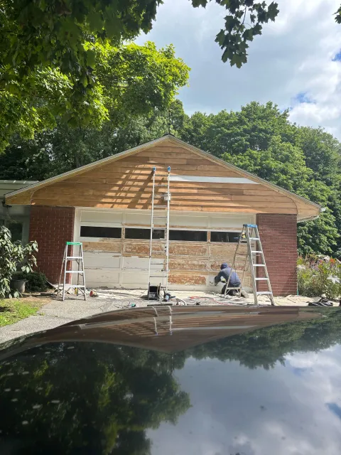 Workers repairing a brick house with a wooden roof frame, reflected in a car hood, under trees
