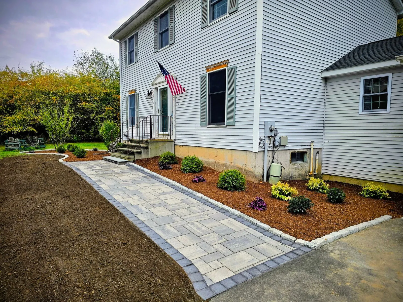A newly installed paver walkway leads to the front door of a white house with a landscaped garden bed.