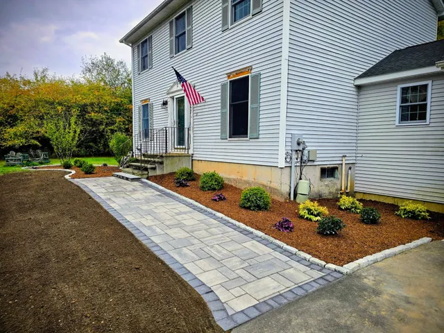 A newly installed paver walkway leads to the front door of a white house with a landscaped garden bed.