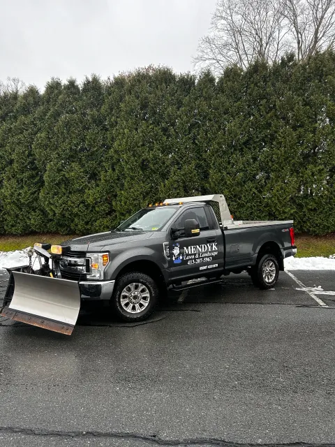 A dark gray pickup truck with a snow plow attached to the front, parked in a lot with some snow on the ground and a row of tall green trees in the background. The truck has branding for 'Mendy Lawn & Landscape' and contact information on the door.