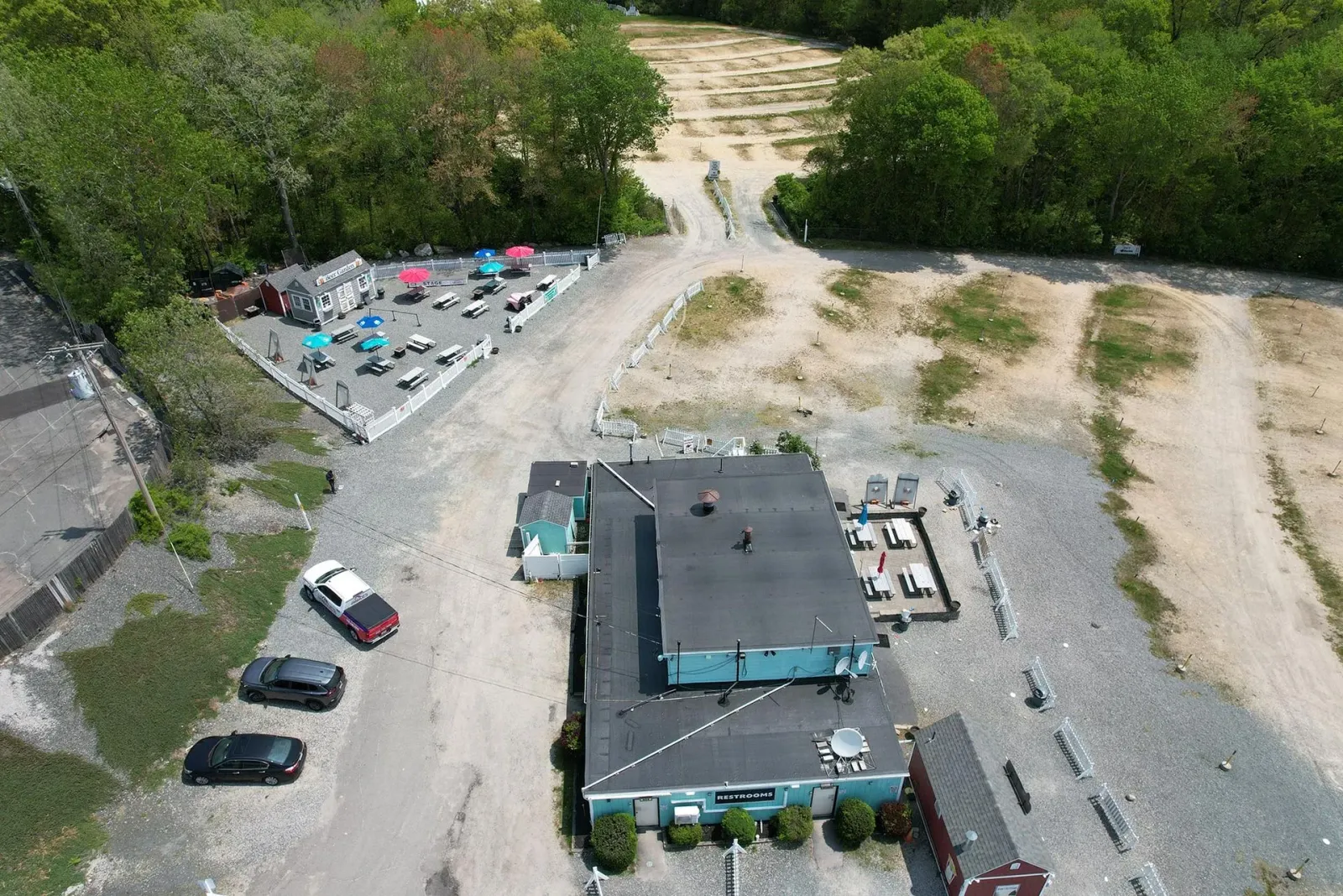 Aerial view of a building, parked cars, and a gravel lot beside trees and a dirt path.
