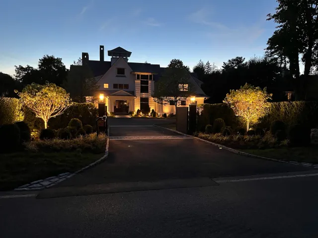A large, multi-story house illuminated by outdoor lighting at dusk, with trees and landscaped bushes in the front yard and a driveway leading up to the house.