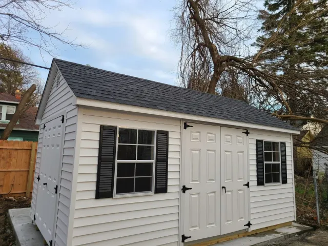 White backyard shed with black shutters and doors, on a concrete foundation, with a peaked black shingle roof, next to a wooden fence and bare trees.