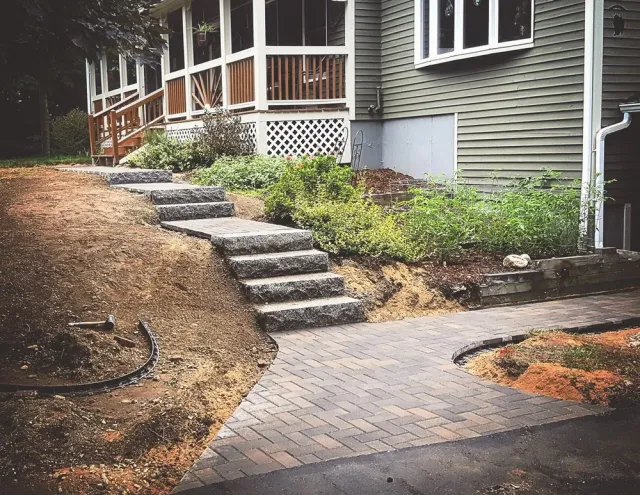 Newly constructed stone steps leading up to the porch of a house with green siding. A paved walkway curves around a garden bed with plants and soil. unfinished landscaping is visible on either side of the walkway.