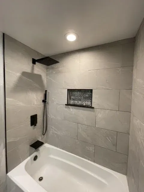 Modern bathroom shower with large gray tiles, a black rainfall showerhead, a handheld shower with a black holder, a built-in niche for toiletries, and a white bathtub.