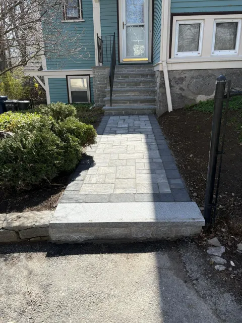 Stone walkway leading to a house entrance, with steps up to the door and bushes along the side.