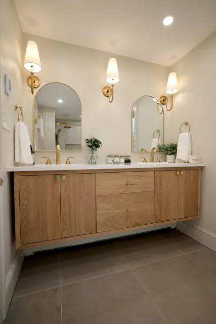 Bathroom vanity with two sinks, wooden cabinet, white countertop, three mirrors, gold fixtures, and wall-mounted lamps.