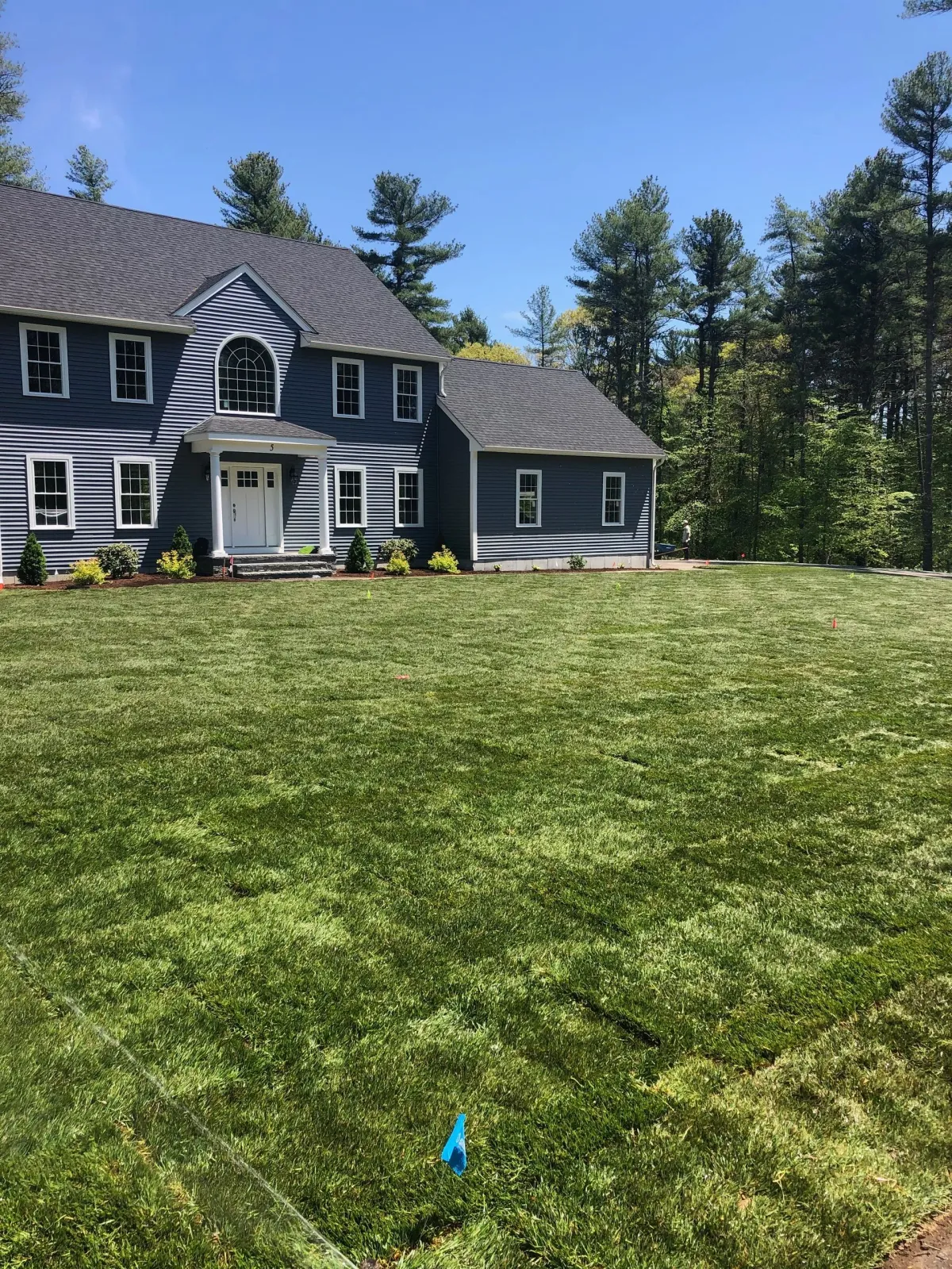 Blue-gray house with white trim beside a large green lawn and trees on a sunny day