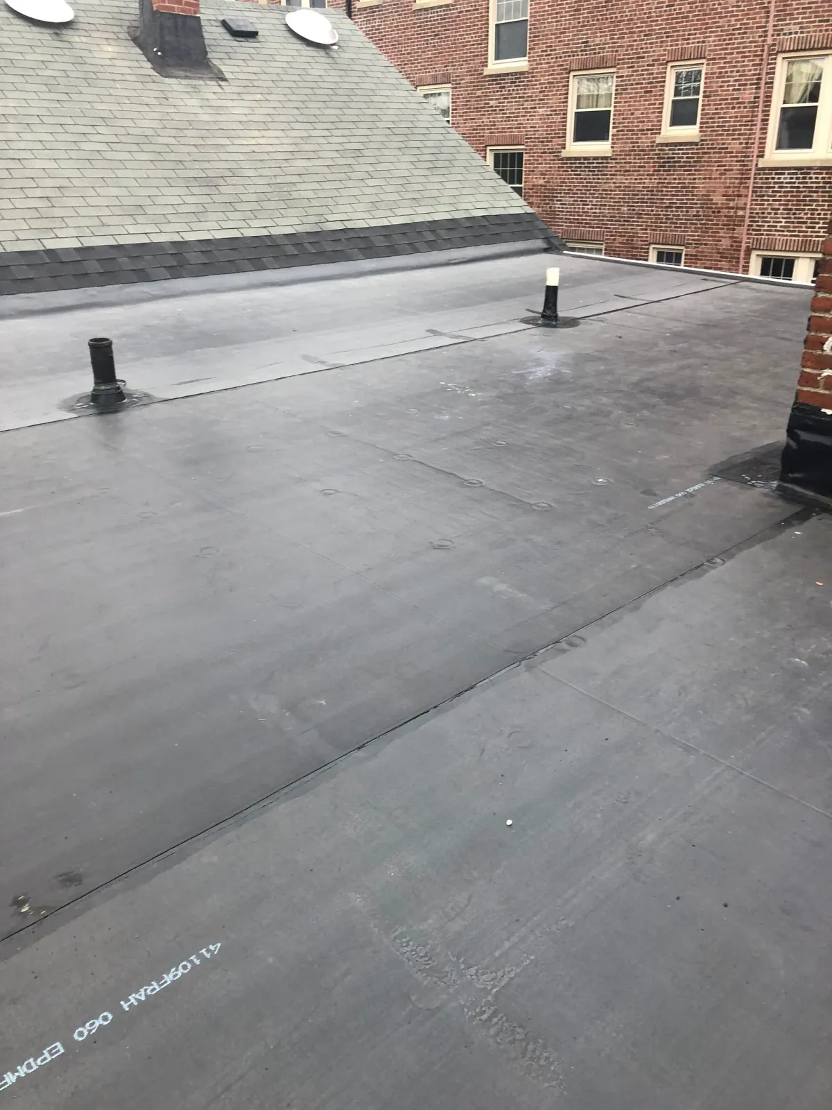 Flat rooftop with black membrane, vents, and adjacent brick buildings under an overcast sky