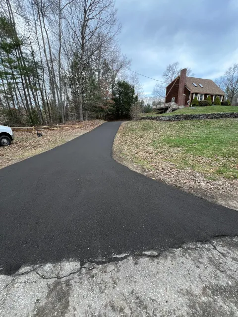 Newly paved asphalt driveway curving toward a house with a brick chimney. Overcast day.