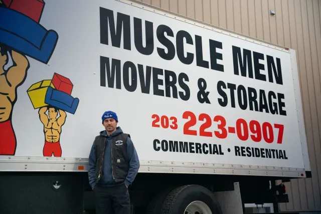 Man standing by Muscle Men Movers truck. Truck has logo and phone number.
