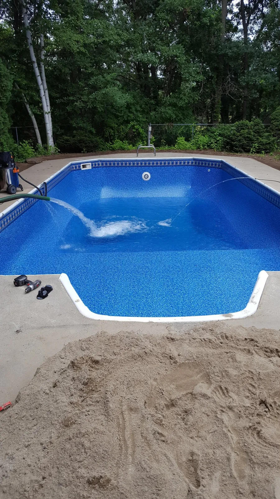 A blue in-ground swimming pool with a concrete deck, surrounded by a wooded backyard. There are tools and equipment on the pool deck, and the pool appears to be under maintenance or cleaning.
