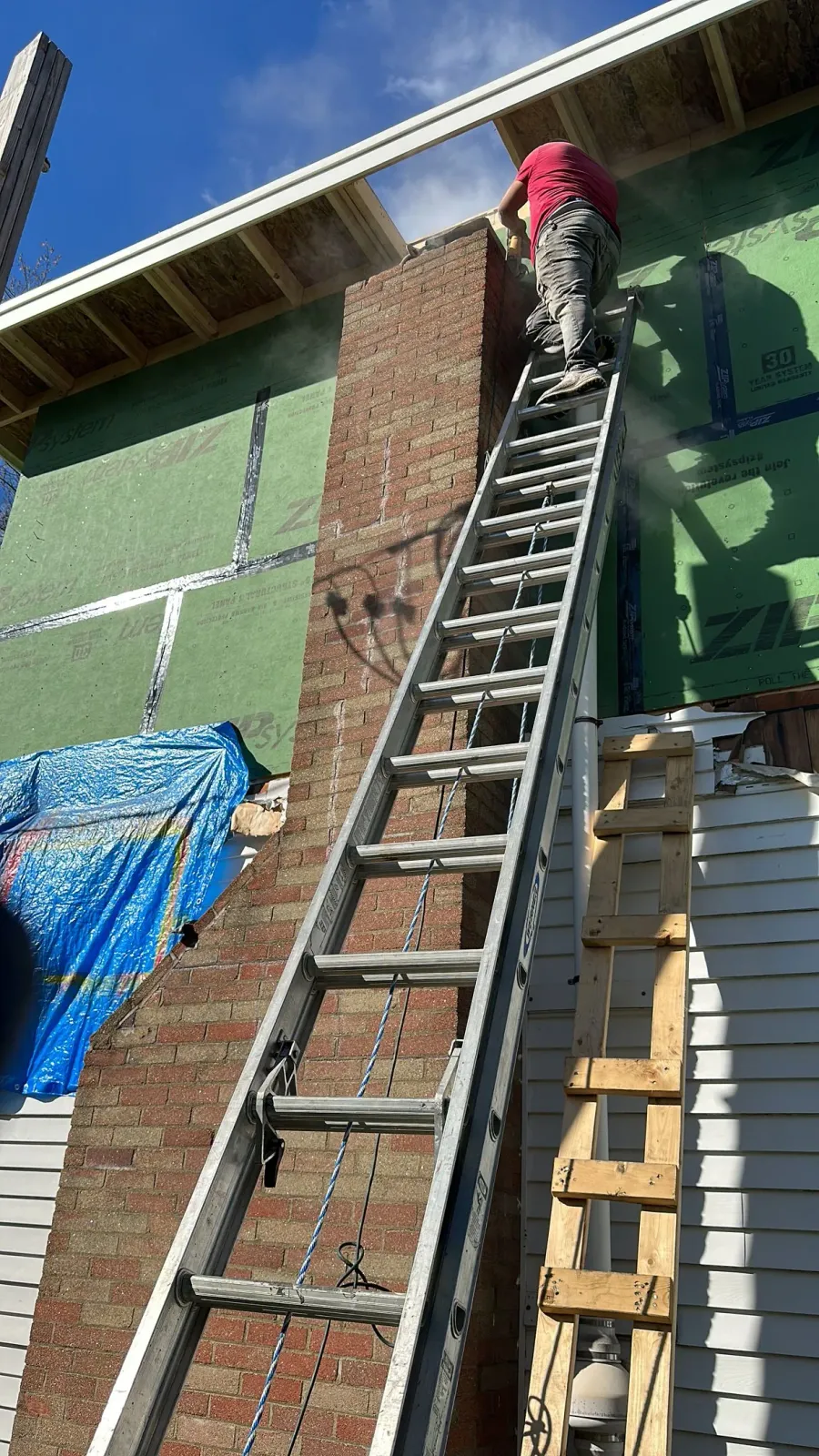 Worker on a ladder painting a brick wall beside a green building.