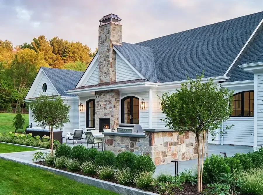 White suburban house with a stone chimney and an outdoor kitchen, patio seating, and a manicured lawn in the foreground.