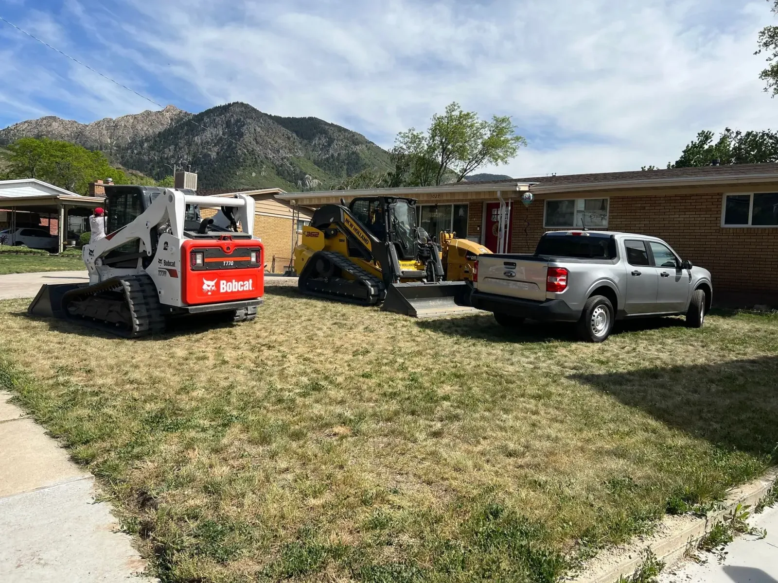 Three construction vehicles and a truck in front of a house with mountains in the background.