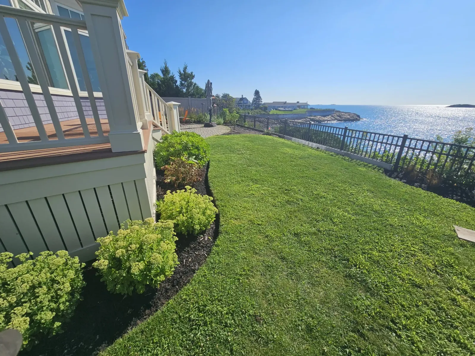 Bright green oceanfront lawn slopes past black mulch beds and a porch toward a black fence and sparkling blue water