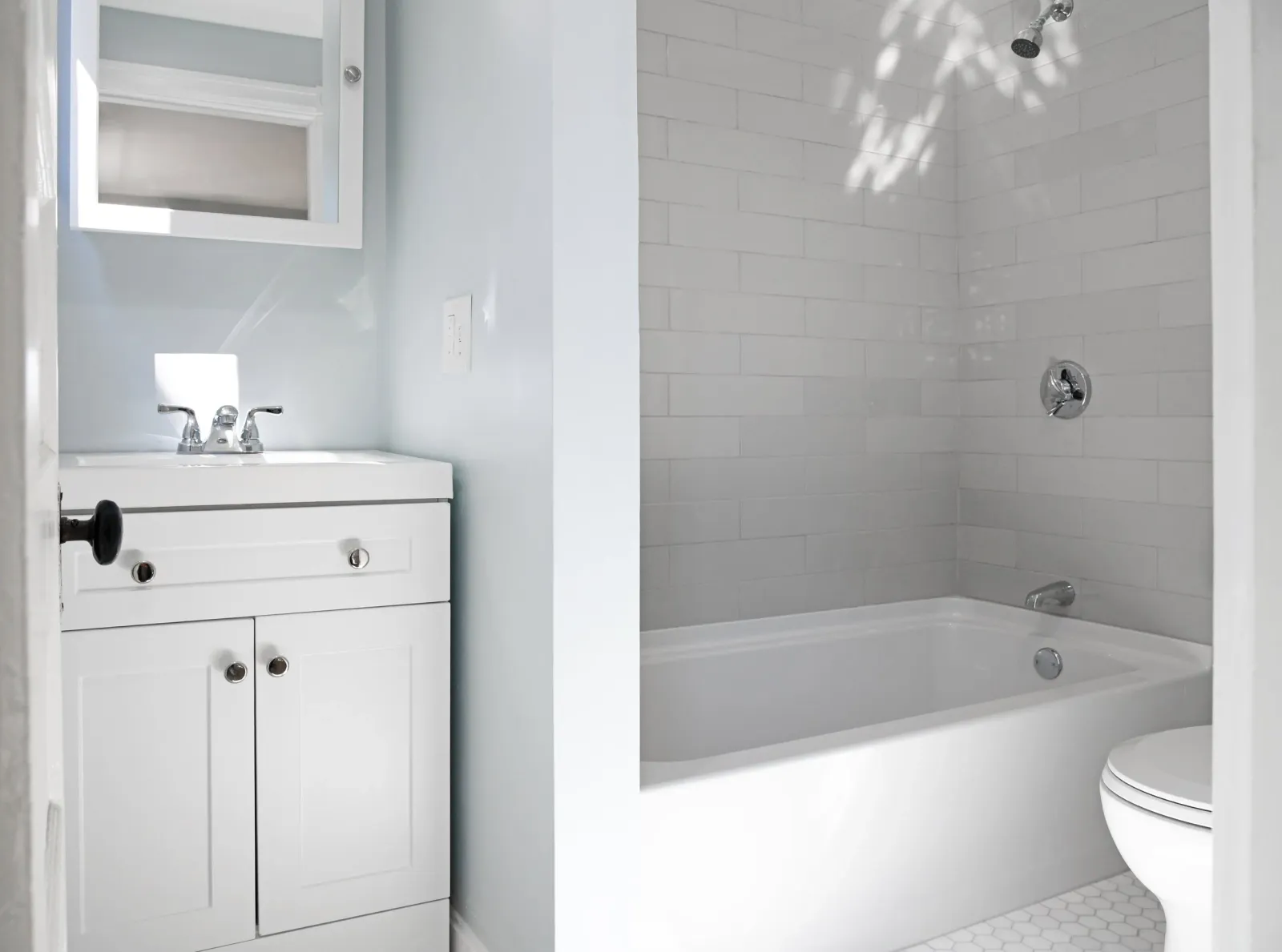 A small bathroom with a white vanity, a mirror cabinet, a bathtub with beige tiles, a showerhead, and a toilet.