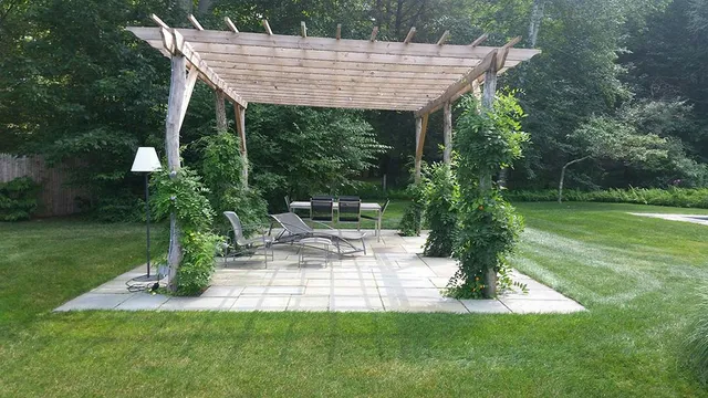 Wooden pergola over a stone patio with outdoor seating, surrounded by green lawn and trees