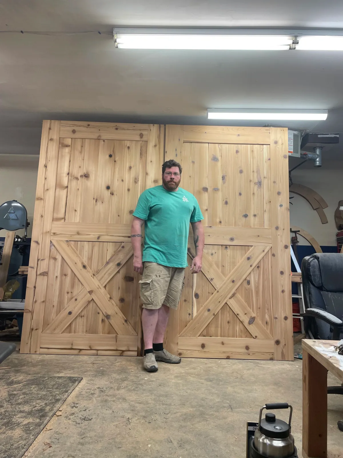 A man standing in a woodworking shop in front of two large wooden barn doors, with tools and workbenches around him.