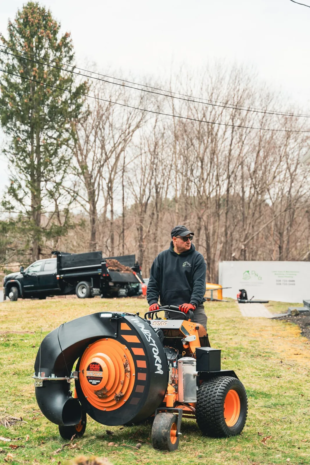 A man in a black hoodie, red gloves, and sunglasses operates a leaf blower machine on a lawn with trees and utility trucks in the background.