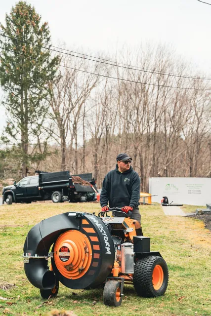 A man in a black hoodie, red gloves, and sunglasses operates a leaf blower machine on a lawn with trees and utility trucks in the background.