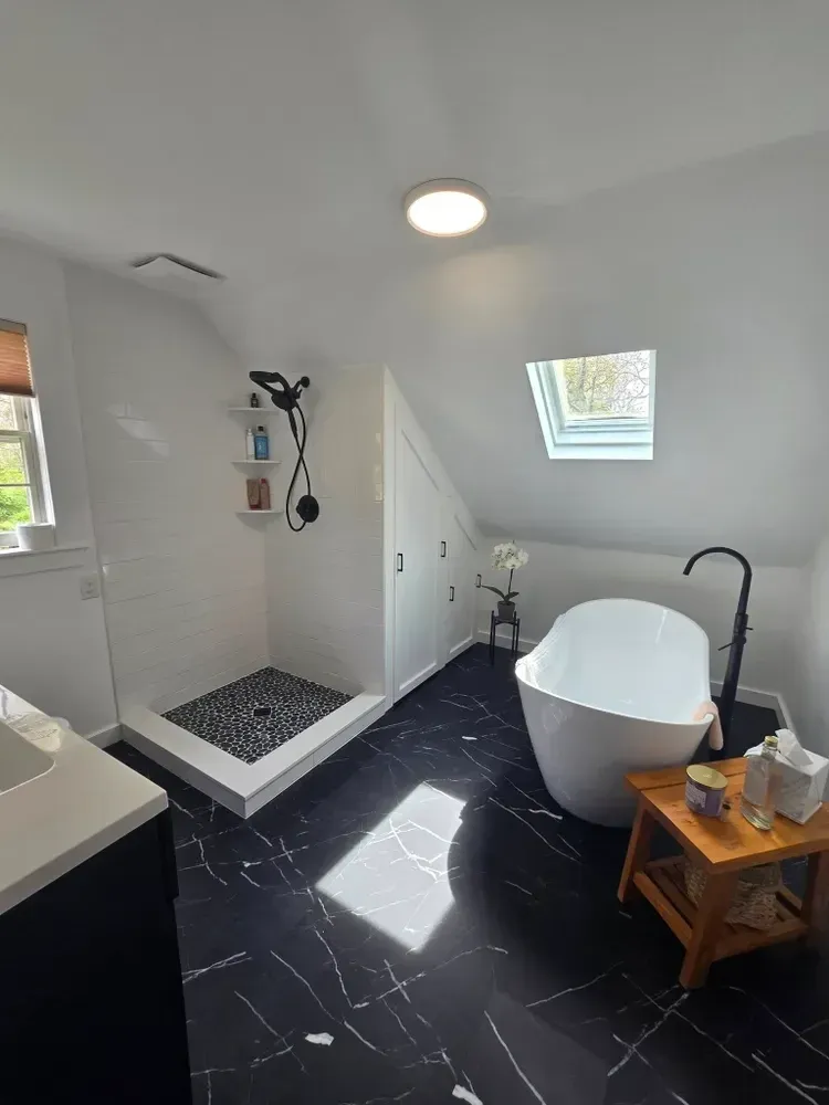 Modern bathroom with a freestanding bathtub, black and white marble floor, skylight, and walk-in shower with black pebble tiles, small black stool with toiletries, and a window with blinds.