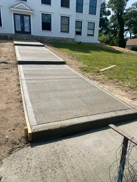 Newly poured concrete sidewalk leading to the entrance of a white house with multiple windows, with wooden forms and some construction materials visible along the sidewalk.
