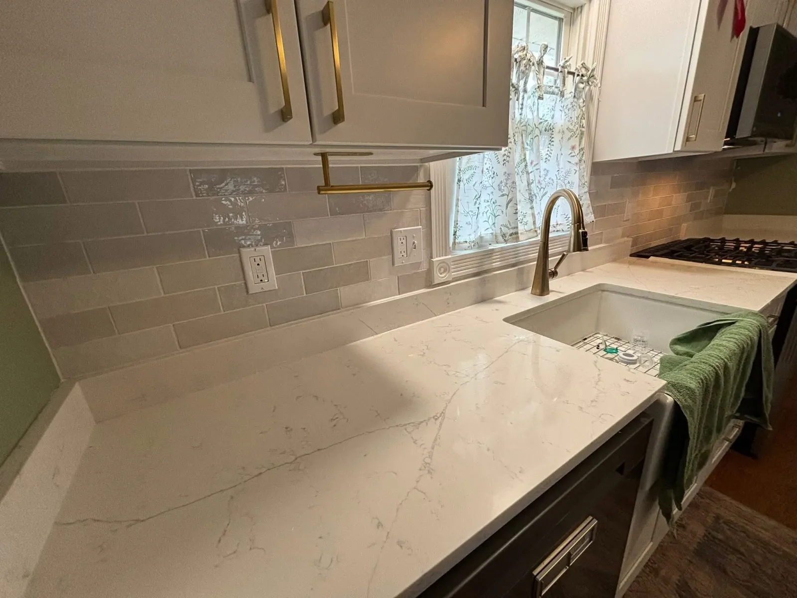 A kitchen with white cabinets, a marble countertop, a brass faucet, a white tile backsplash, a window with floral curtains, and a gas stove.