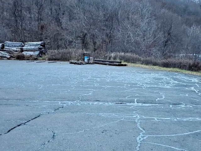A paved lot with cracks, featuring stacked logs on the left and a small blue structure in the background against woods.