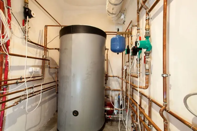 Utility room with a large gray water heater, copper pipes, and valves along narrow white walls.