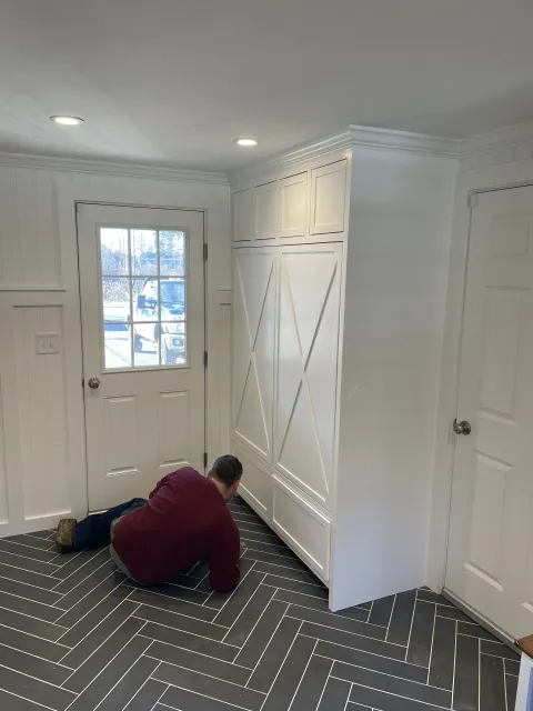 Man kneeling on a herringbone patterned black and white tile floor in front of white paneled walls, a door with a window, and a built-in cabinet or closet with crisscrossed panel doors.