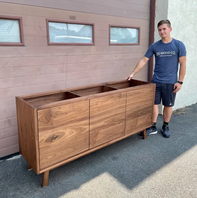Young man standing outside next to a mid-century modern wooden sideboard with three cabinet doors and open storage compartments, against a pink wooden wall and concrete ground.