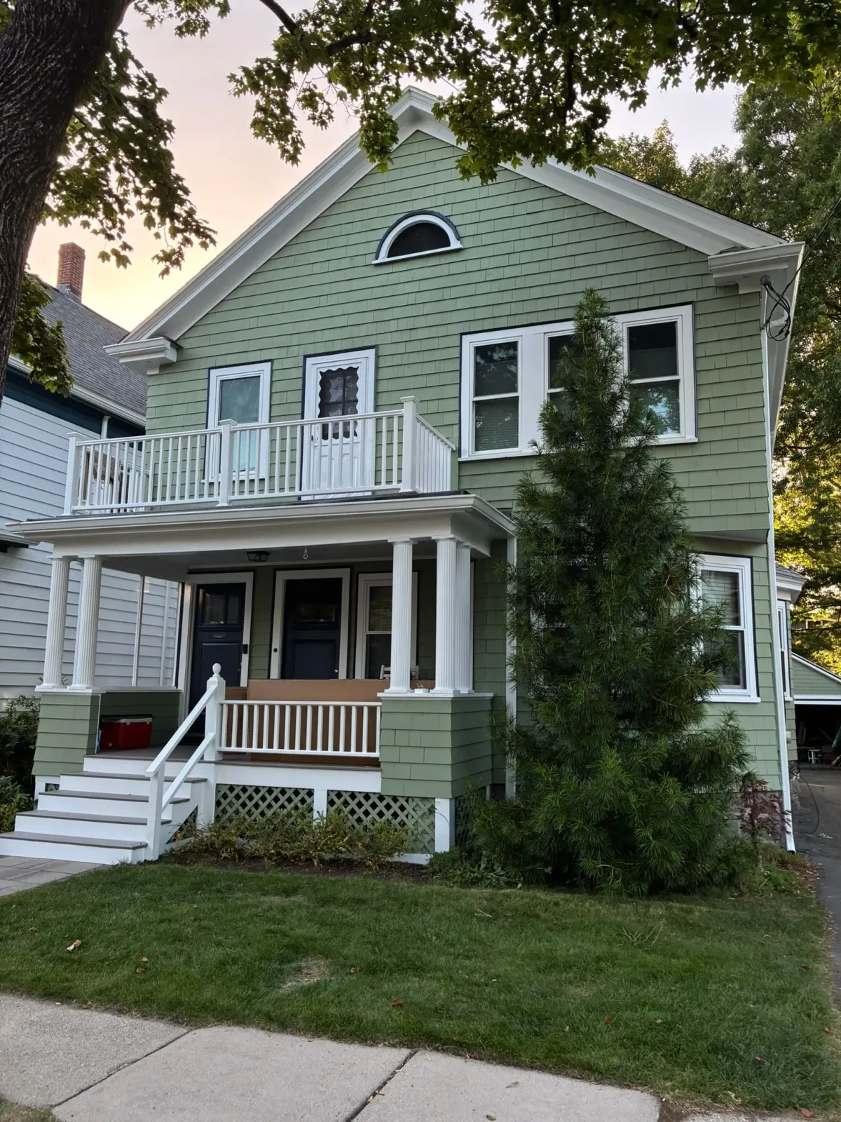 Two-story green house with white trim, front porch, balcony, and steps under leafy trees