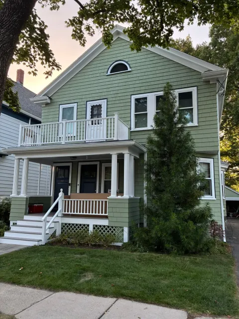 Two-story green house with white trim, front porch, balcony, and steps under leafy trees