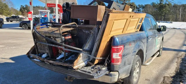 Blue pickup truck with a damaged bed piled with debris, parked on a snowy roadside outdoors